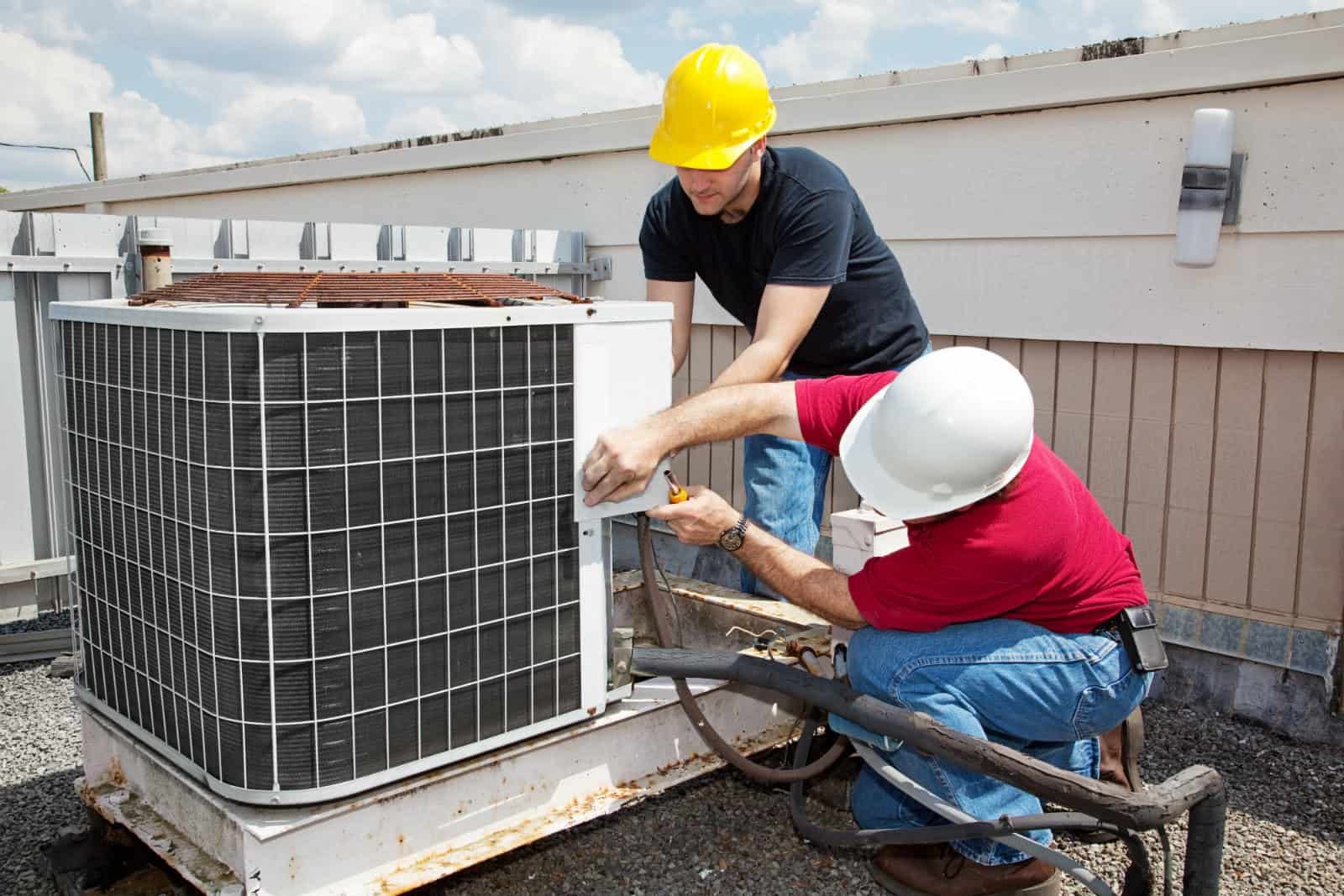 Two HVAC technicians wearing safety helmets perform maintenance on a rooftop air conditioning unit, using tools to inspect and repair the system.