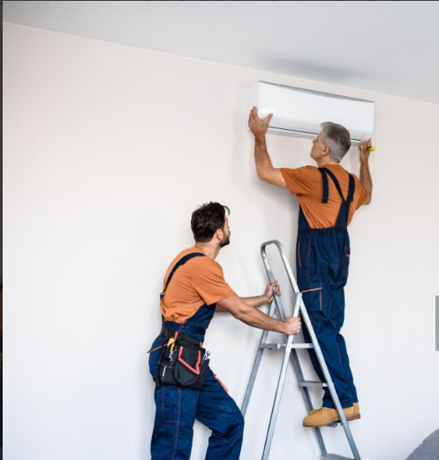 Two technicians performing air conditioning installation, with one standing on a ladder mounting a split AC unit and the other assisting from the ground.